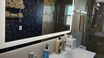 Une salle de bain dotée d’un grand miroir, d’un lavabo blanc, d’un distributeur de savon manuel et d’une cabine de douche en verre.