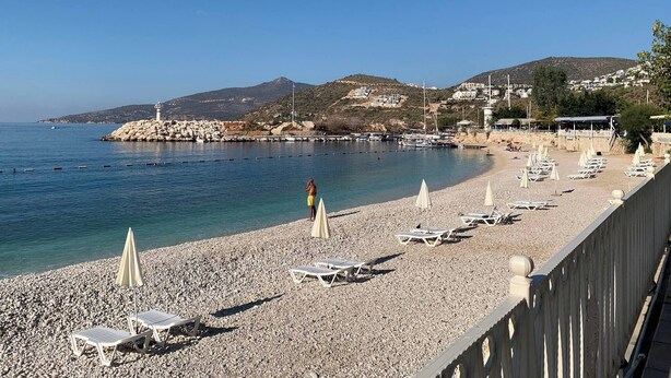 A pebble beach with lounge chairs and umbrellas, a lighthouse, and buildings in the background. - Sylvana Apartment- a Little Piece of Kalkan Heaven (Kaş)