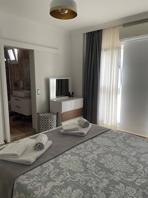 A hotel room with a bed, a bedside table, a mirror, a window with curtains, and a bathroom visible through an open door. - Sylvana Apartment- a Little Piece of Kalkan Heaven (Kaş)