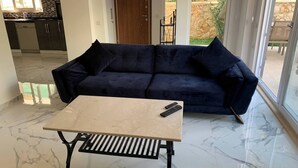 A living room with a dark blue sofa, a small beige table, and a kitchen in the background. - Sylvana Apartment- a Little Piece of Kalkan Heaven (Kaş)