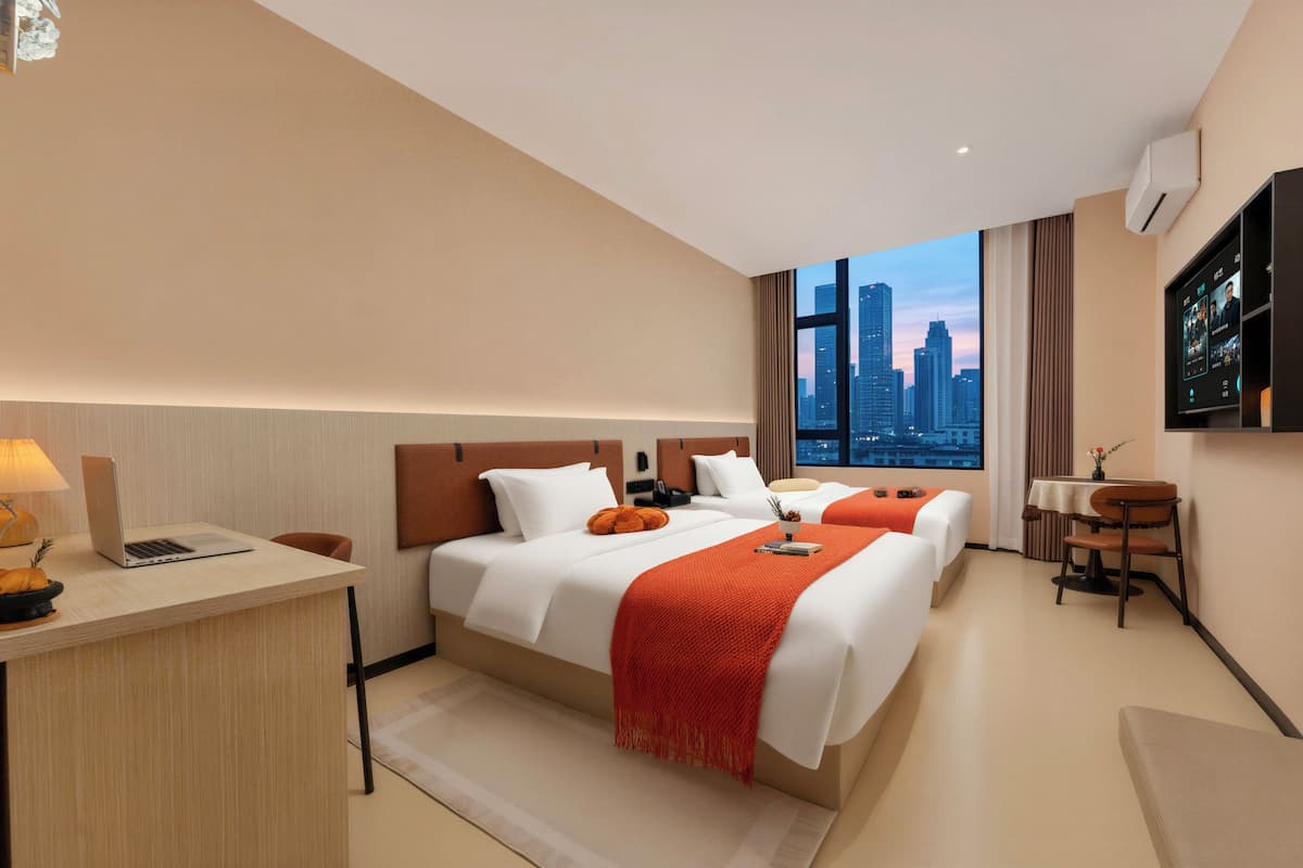 A hotel room with a city view, a desk with a laptop, a bed with white bedding and orange accents, and a TV mounted on the wall.