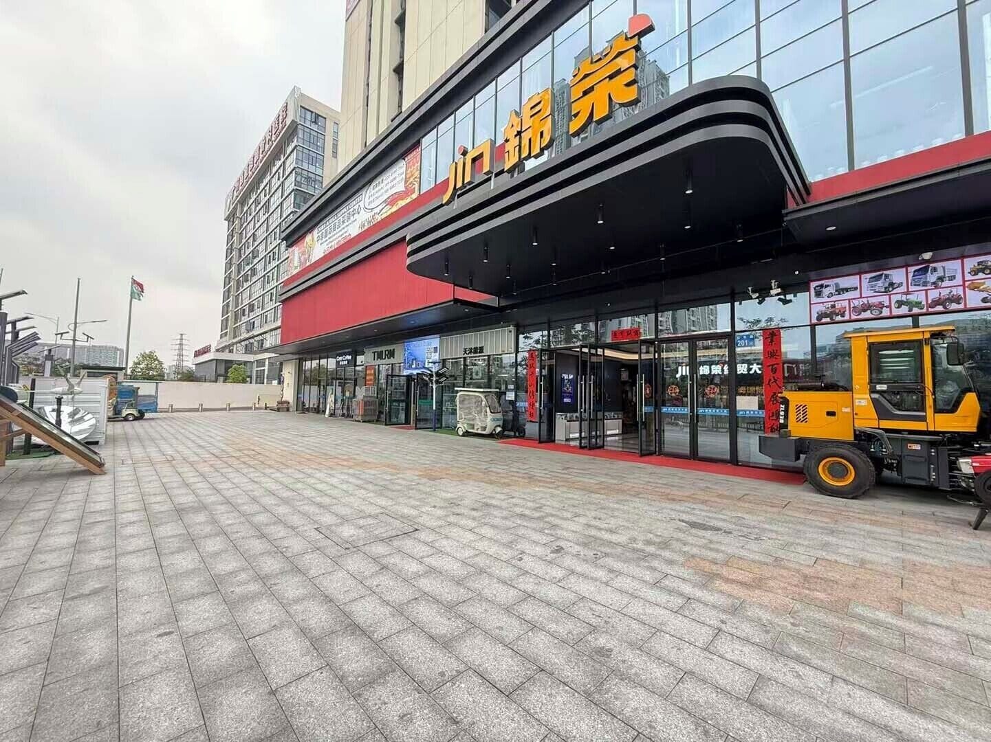 Un immeuble commercial doté d’une grande façade vitrée, d’un auvent rouge et noir, et d’un chariot élévateur jaune garé à l’extérieur.