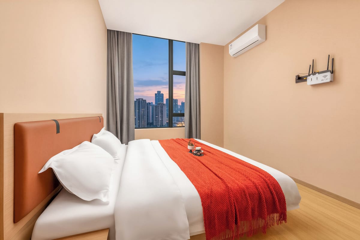 A hotel room with a large window, a bed with white bedding and a red throw, and a wall-mounted air conditioning unit.