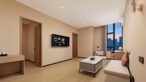 Una habitación de hotel moderna con una televisión de pantalla plana, una zona de estar con sofá y reposapiés, una mesita y una ventana con vista a la ciudad.