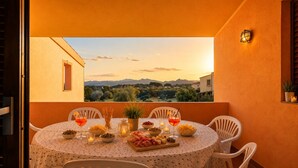 Un tavolo rotondo con sedie bianche su un balcone esterno, apparecchiato con cibo, bevande e candele, con vista su un paesaggio suggestivo.