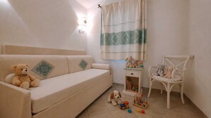 Et værelse med en beige sofa, et lille hvidt bord med legetøj, en hvid stol og et vindue med gardin.