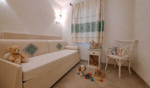 A room with a beige sofa, a small white table with toys, a white chair, and a window with a curtain. - Porto San Paolo - Sea View House with Garden (Porto San Paolo)