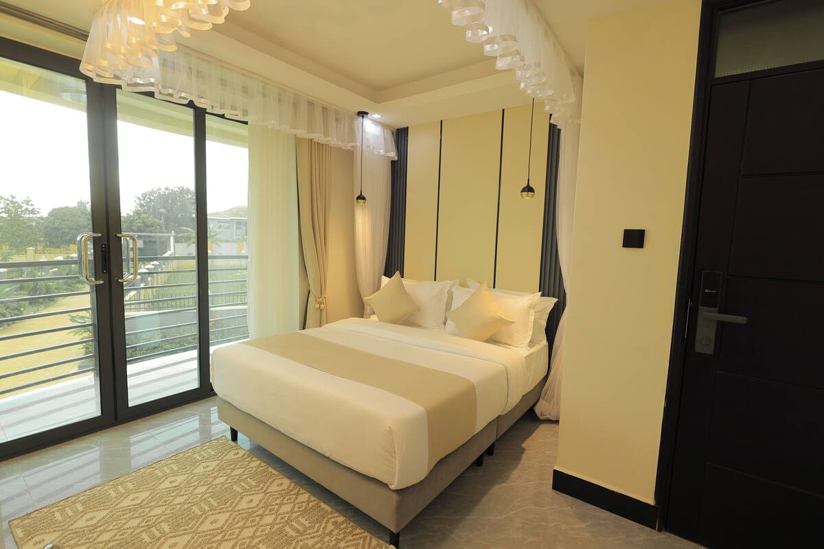 A bedroom with a large bed, a balcony with a railing, and a view of greenery.