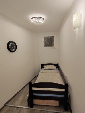 Um quarto pequeno e estreito com uma cama de solteiro, um relógio na parede, uma janela com desenho decorativo e um lustre no teto.