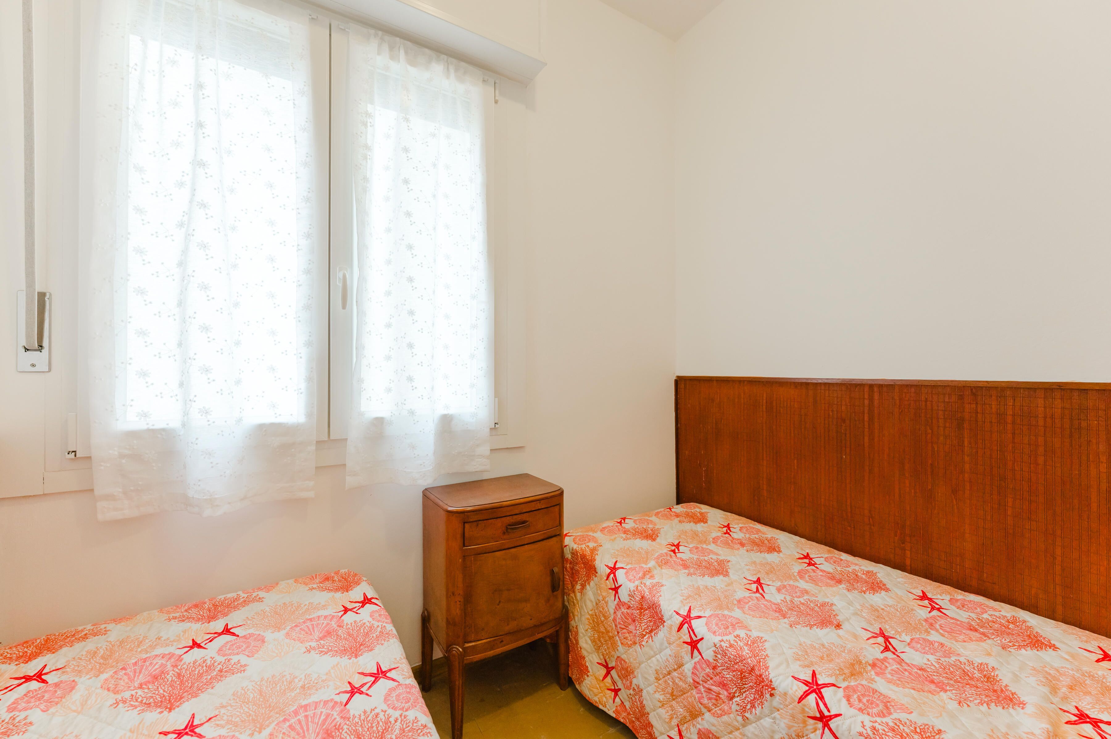 A bedroom with two single beds, a wooden nightstand, and a window with sheer curtains.