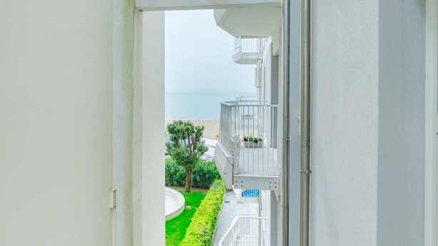 A balcony with a view of the sea, a green lawn, and a white building. - Anna Flat (Lido di Jesolo)