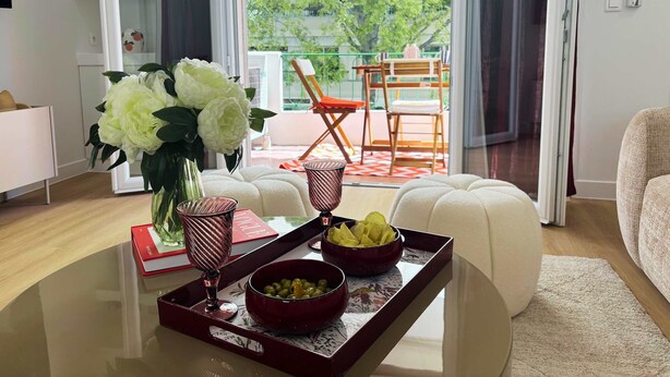 Een moderne woonkamer met een glazen salontafel, een vaas met bloemen, een dienblad met schalen en uitzicht op een balkon met buitenmeubilair.