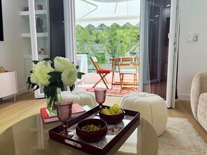 A modern living room with a glass coffee table, a vase with flowers, a tray with bowls, and a view of a balcony with outdoor furniture. - Barbara – 2BDR, 2 Bathrooms by the Beach (Cannes)