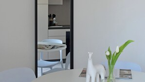 Una mesa redonda blanca con un florero y una figurita blanca. Un espejo refleja una cocina con armarios blancos y una silla blanca.