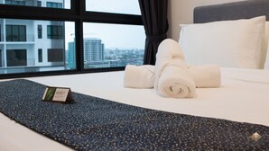 Habitación de hotel con una cama, una toalla enrollada, un almohadón y una ventana con vista a la ciudad.