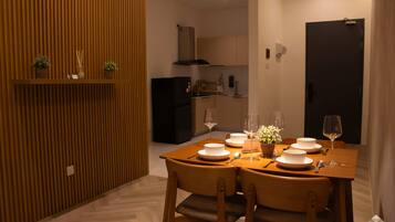 A dining area with a wooden table set for four, surrounded by chairs, with a kitchen in the background.