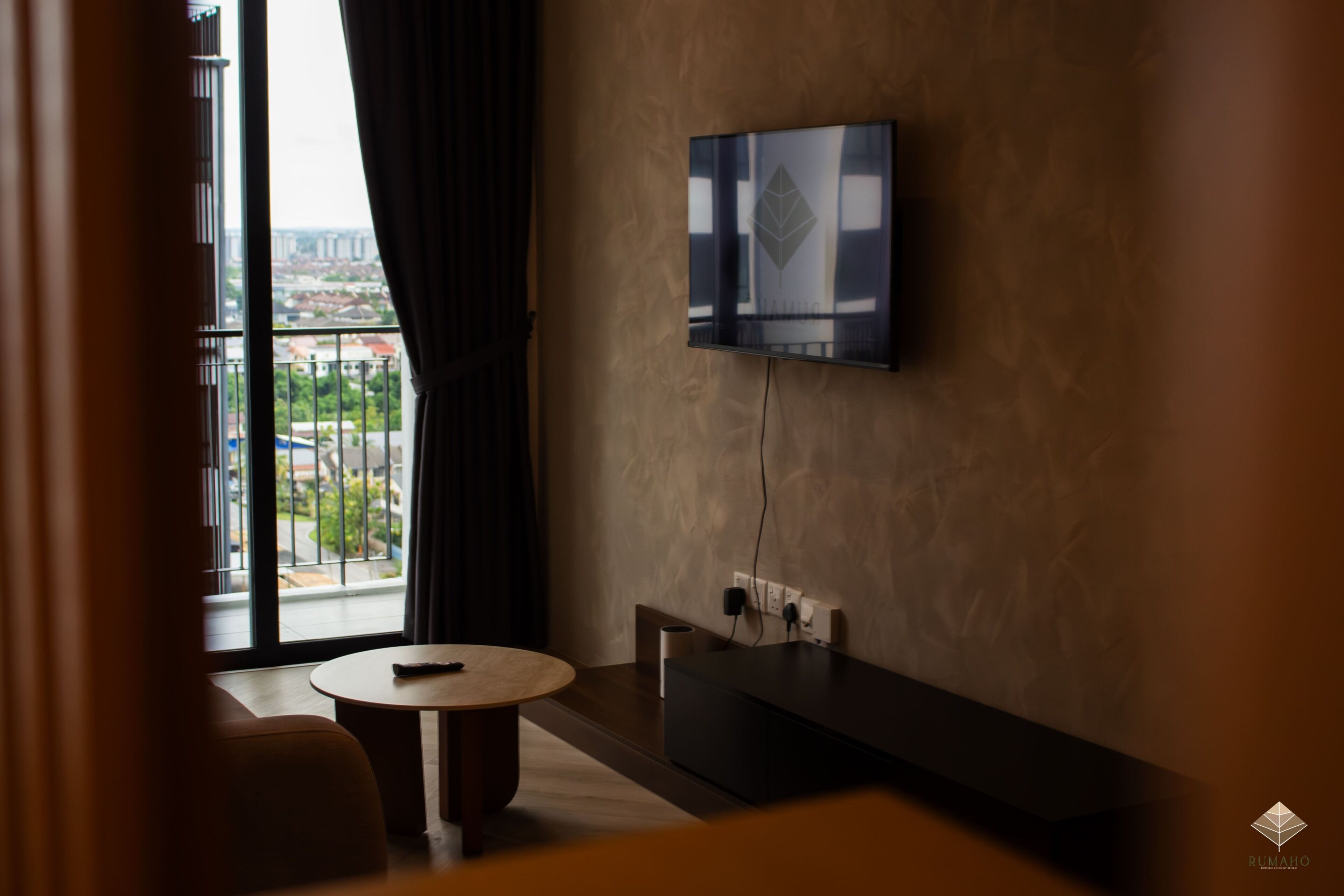 Habitación de hotel con televisor montado en la pared, una mesa redonda, un sofá y un balcón con vistas a edificios.