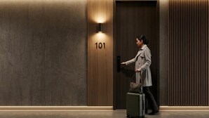 A person entering a room with a wooden door marked '101', a wall-mounted light, and a suitcase.
