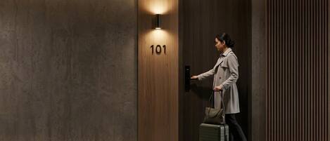 A person entering a room with a wooden door marked '101', a wall-mounted light, and a suitcase.