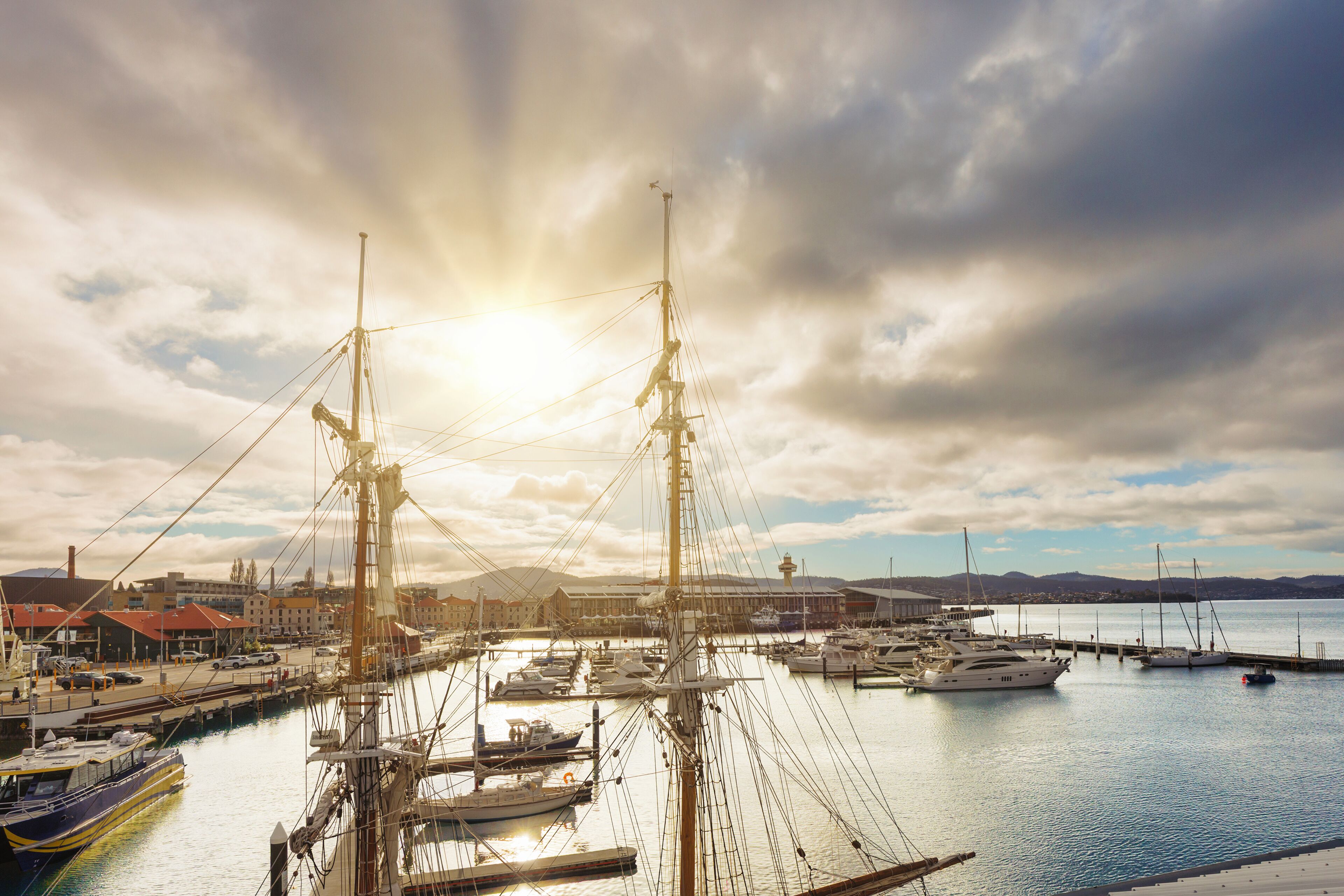 A marina with multiple boats docked, a prominent sailboat in the foreground, and a backdrop of buildings and hills.