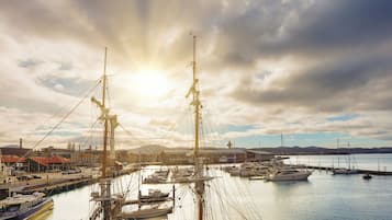 A marina with multiple boats docked, a prominent sailboat in the foreground, and a backdrop of buildings and hills.