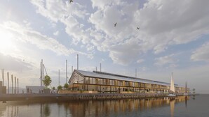 A waterfront building with a modern design, featuring a long structure with a sloped roof, large windows, and a reflective water surface in the foreground.