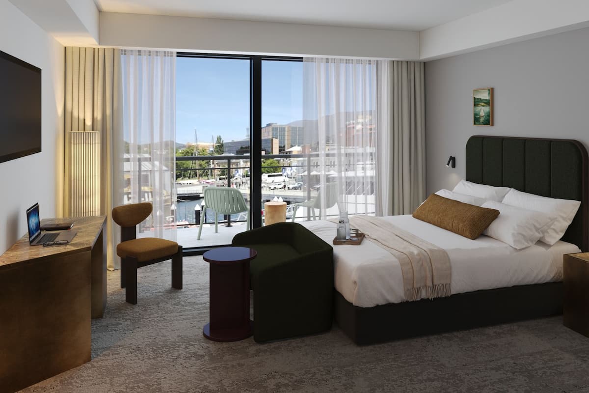 Ein modernes Hotelzimmer mit einem großen Bett, einem Schreibtisch mit einem Laptop, einem Sessel und Blick auf die Stadt durch große Fenster.