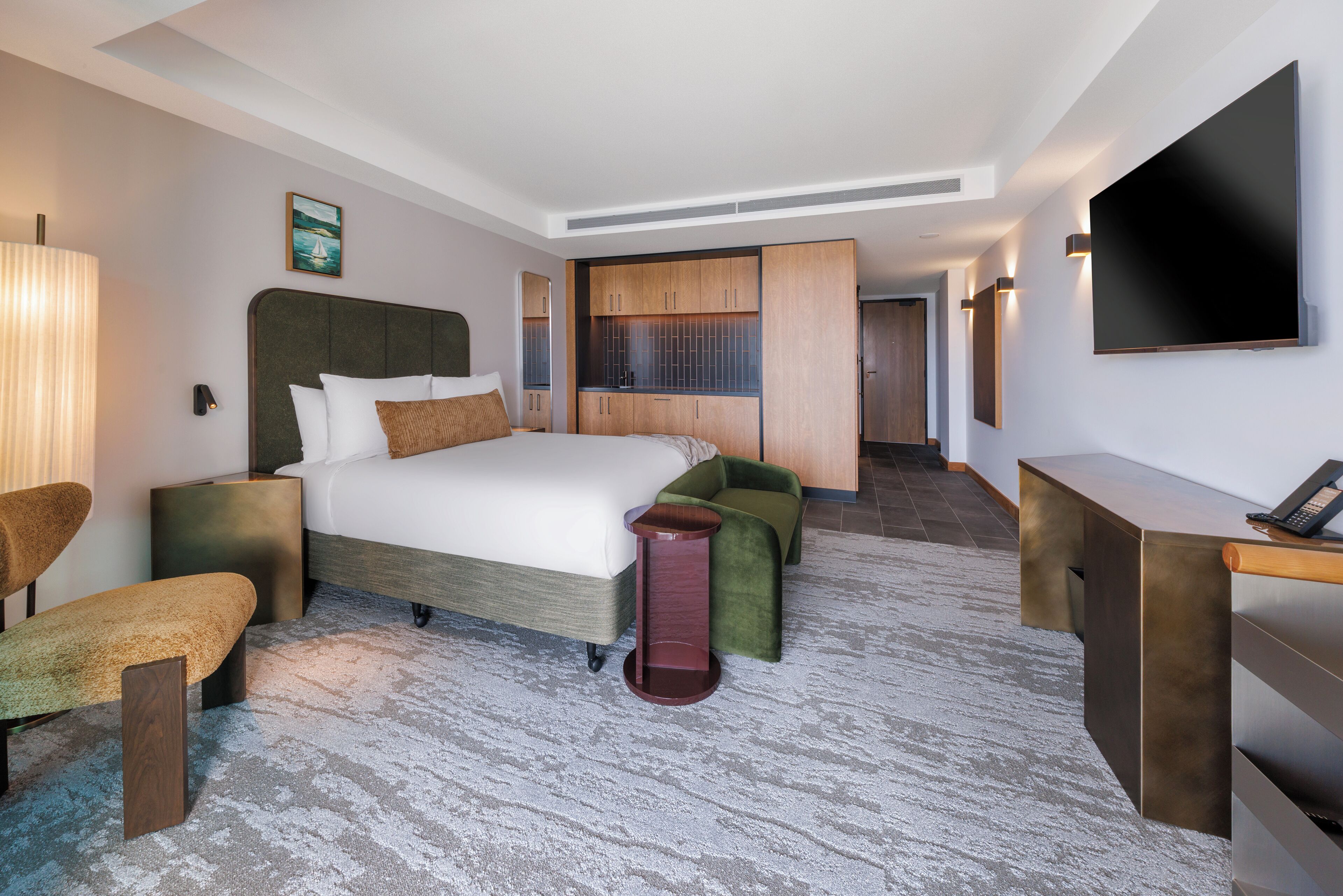 A hotel room with a bed, bedside tables, a desk, a chair, a TV, and a view of the hallway.