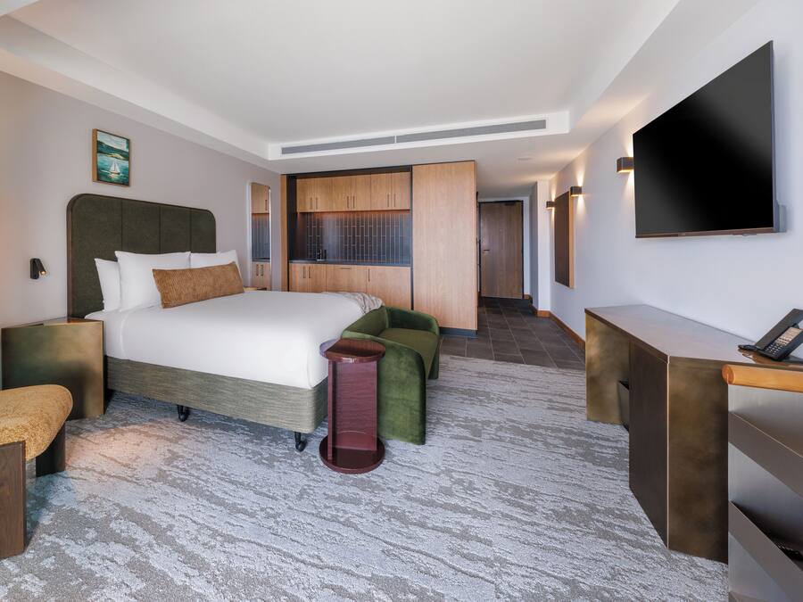 A hotel room with a bed, bedside tables, a desk, a chair, a TV, and a view of the hallway.