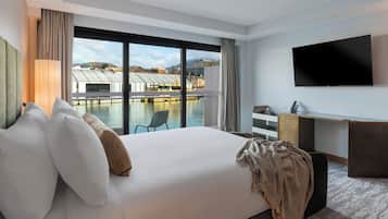 A hotel room with a large bed, a desk, a TV, and a view of a body of water with boats.