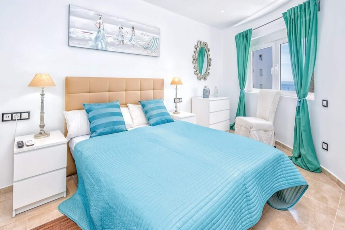 Un dormitorio con una cama grande, dos mesitas de noche, una mesita auxiliar, una silla, un ventanal con cortinas y una obra de arte colgada en la pared.