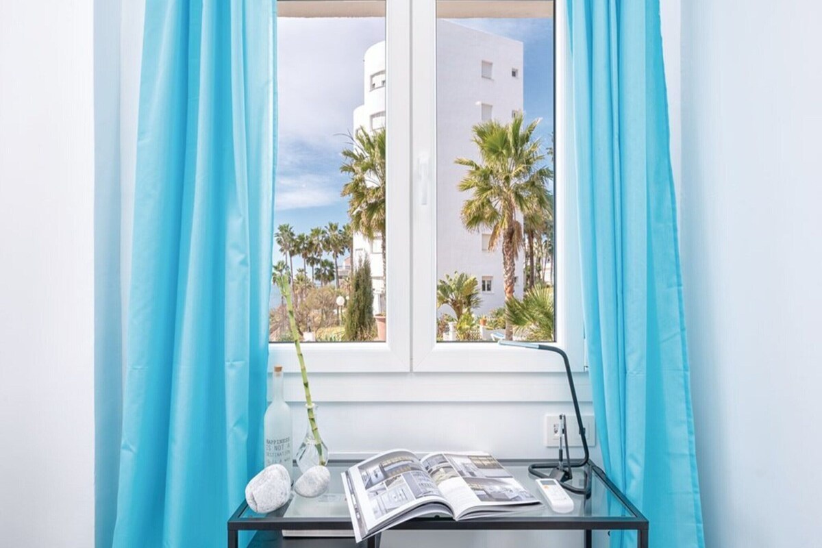 Una habitación con ventana, un escritorio con una revista, una lámpara y un jarrón con planta.