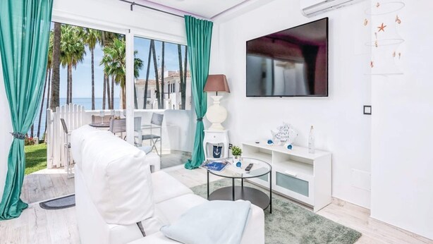Un soggiorno moderno con un divano bianco, un tavolino da caffè in vetro, una TV a parete e vista su palme e mare attraverso ampie finestre.
