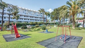 Un’area giochi con uno scivolo rosso e altalene, circondata da erba e palme, con un edificio bianco sullo sfondo.