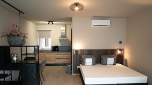 Een moderne slaapkamer met een bed, nachtkastjes, een bureau en een stoel. Achtergrond toont een keuken met een raam.