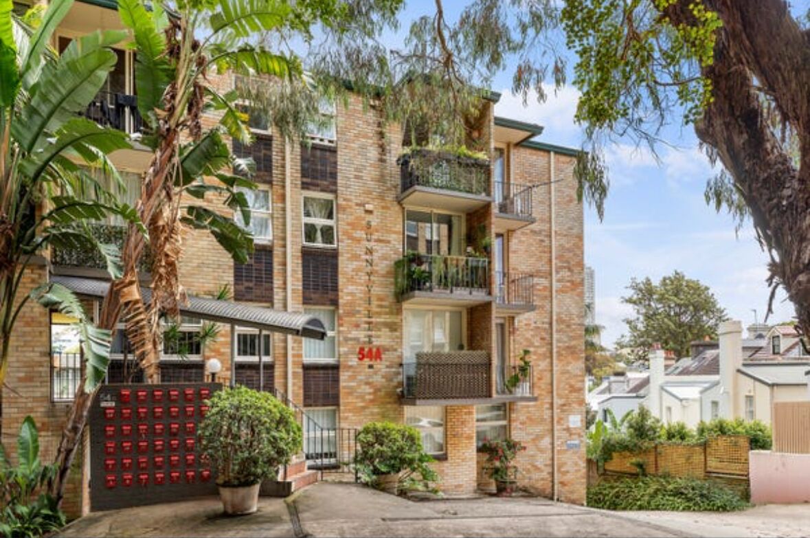 Un immeuble résidentiel à plusieurs étages, doté de balcons, d’une façade en briques et entouré de verdure.