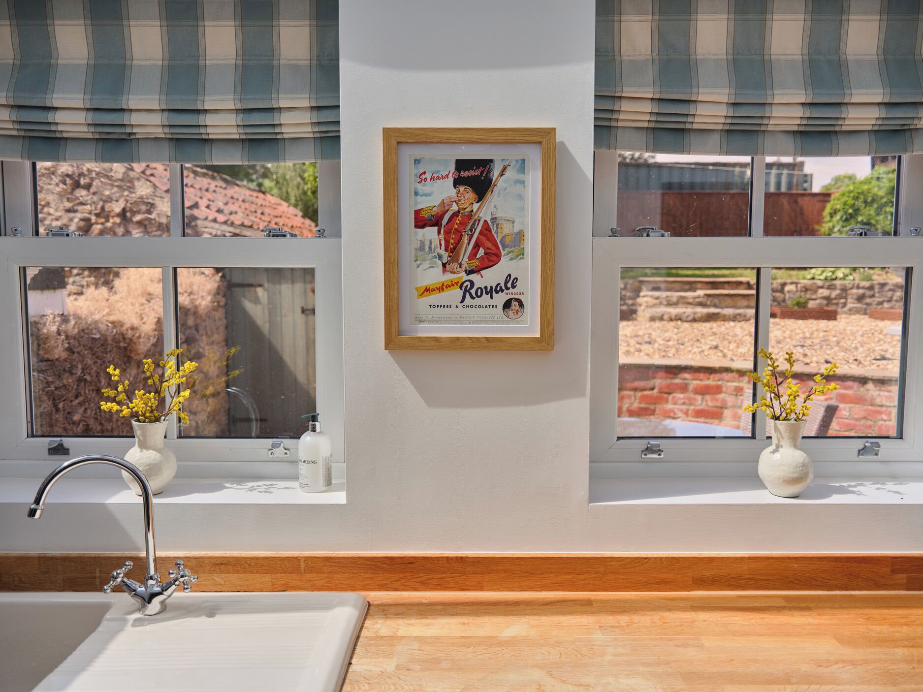 Un baño con un lavamanos, dos jarrones con flores amarillas, un anuncio vintage enmarcado y una ventana que da a un muro de ladrillo y vegetación.