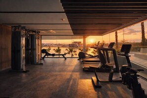 A gym with exercise equipment and a large window overlooking a cityscape at sunset. - The Aqua Luxury Apart Hotel Waterfront (Choeng Thale)