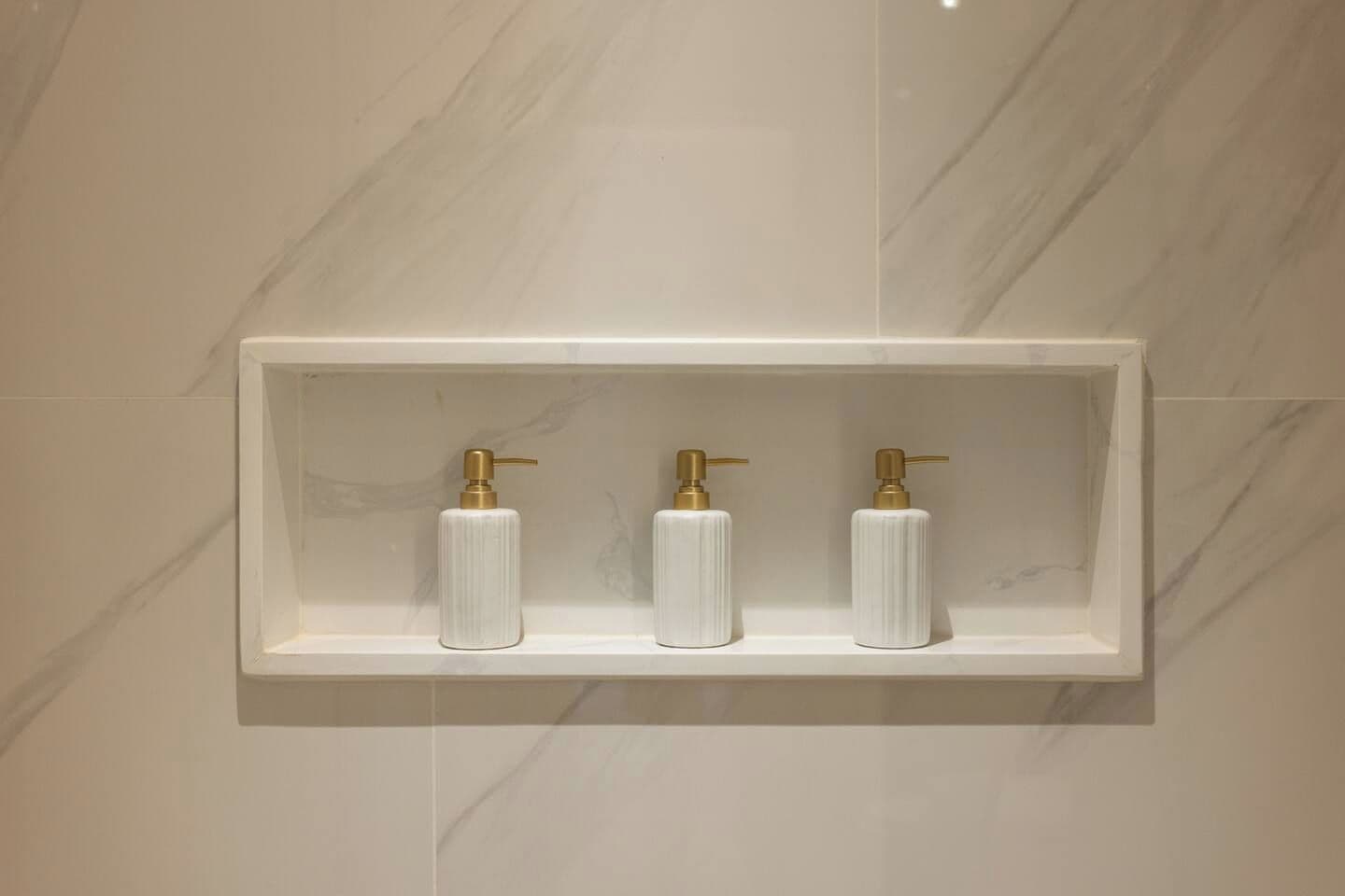 Une salle de bain avec un mur en marbre et une étagère blanche sur laquelle sont posés trois distributeurs de savon blancs, surmontés d’un couvercle doré.