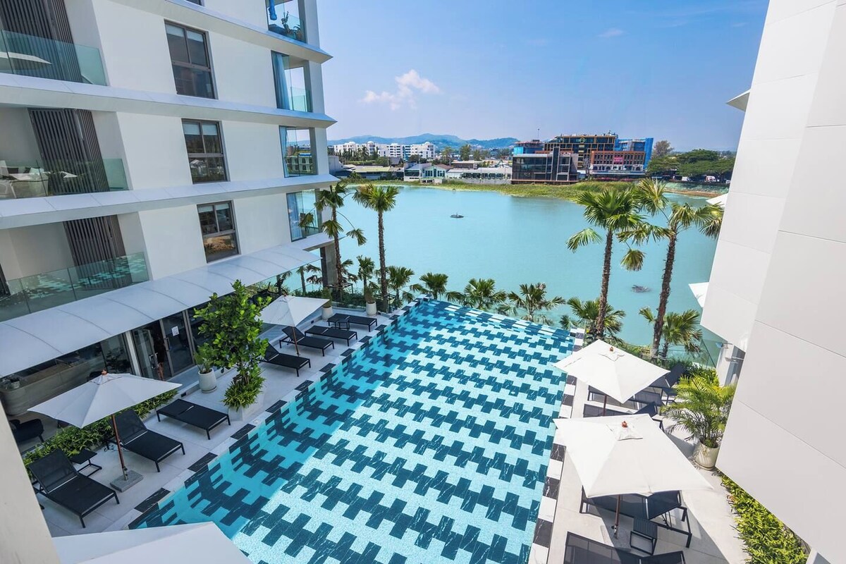 A rooftop pool with a geometric tile pattern, surrounded by lounge chairs and umbrellas, overlooking a body of water and buildings.