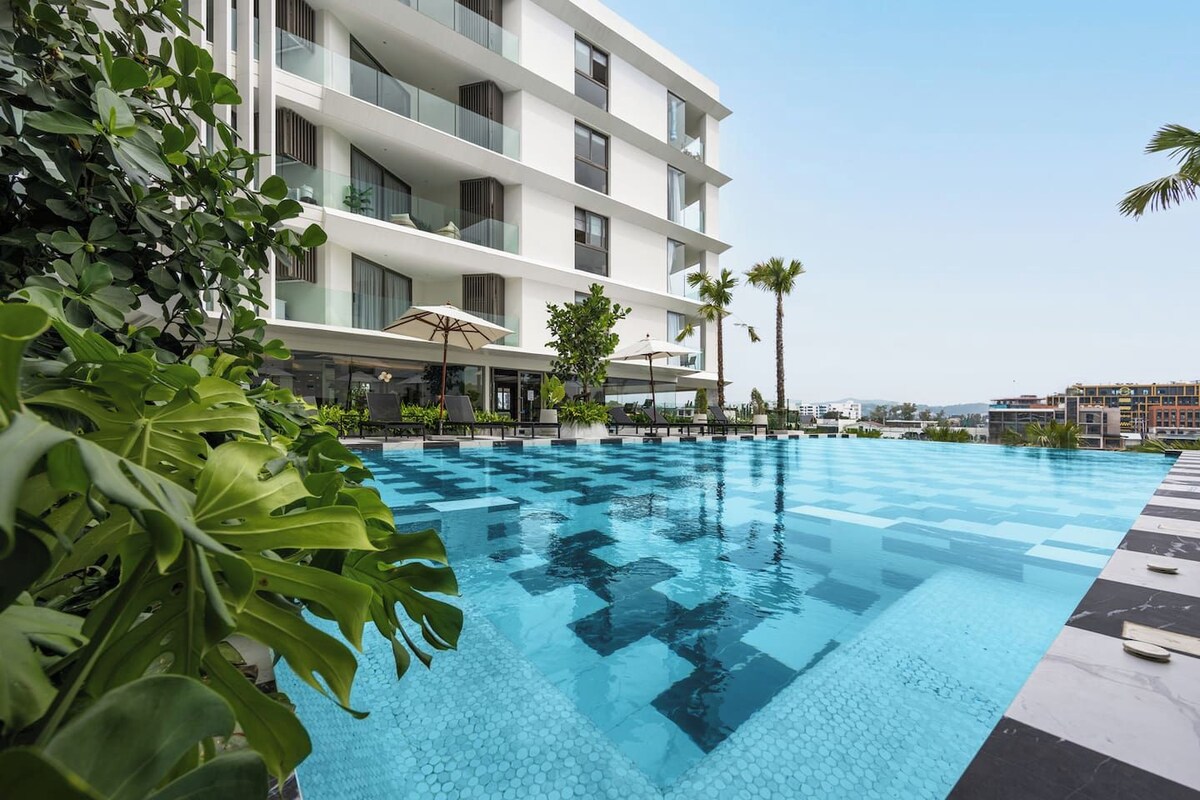 A swimming pool with a checkered pattern, surrounded by lounge chairs and umbrellas, with a multi-story building in the background.