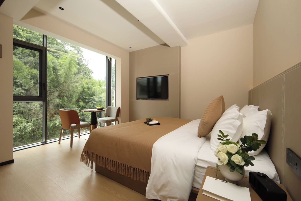 Ein modernes Hotelzimmer mit einem großen Bett, einem Fernseher, einem Schreibtisch mit Stuhl und Blick auf Grünfläche durch das Fenster.