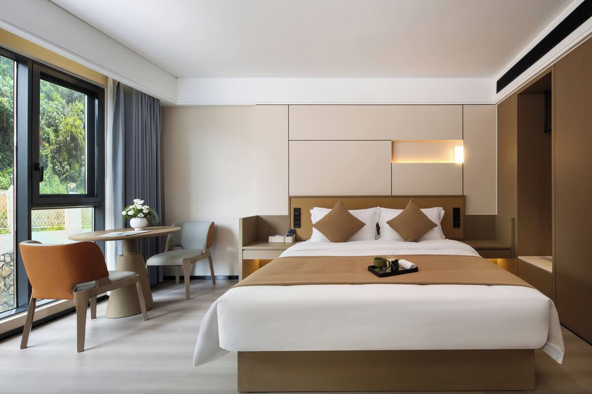 Ein modernes Hotelzimmer mit einem großen Bett, einem Schreibtisch mit Stuhl, einem kleinen Tisch mit einer Vase und Blick durch das Fenster.