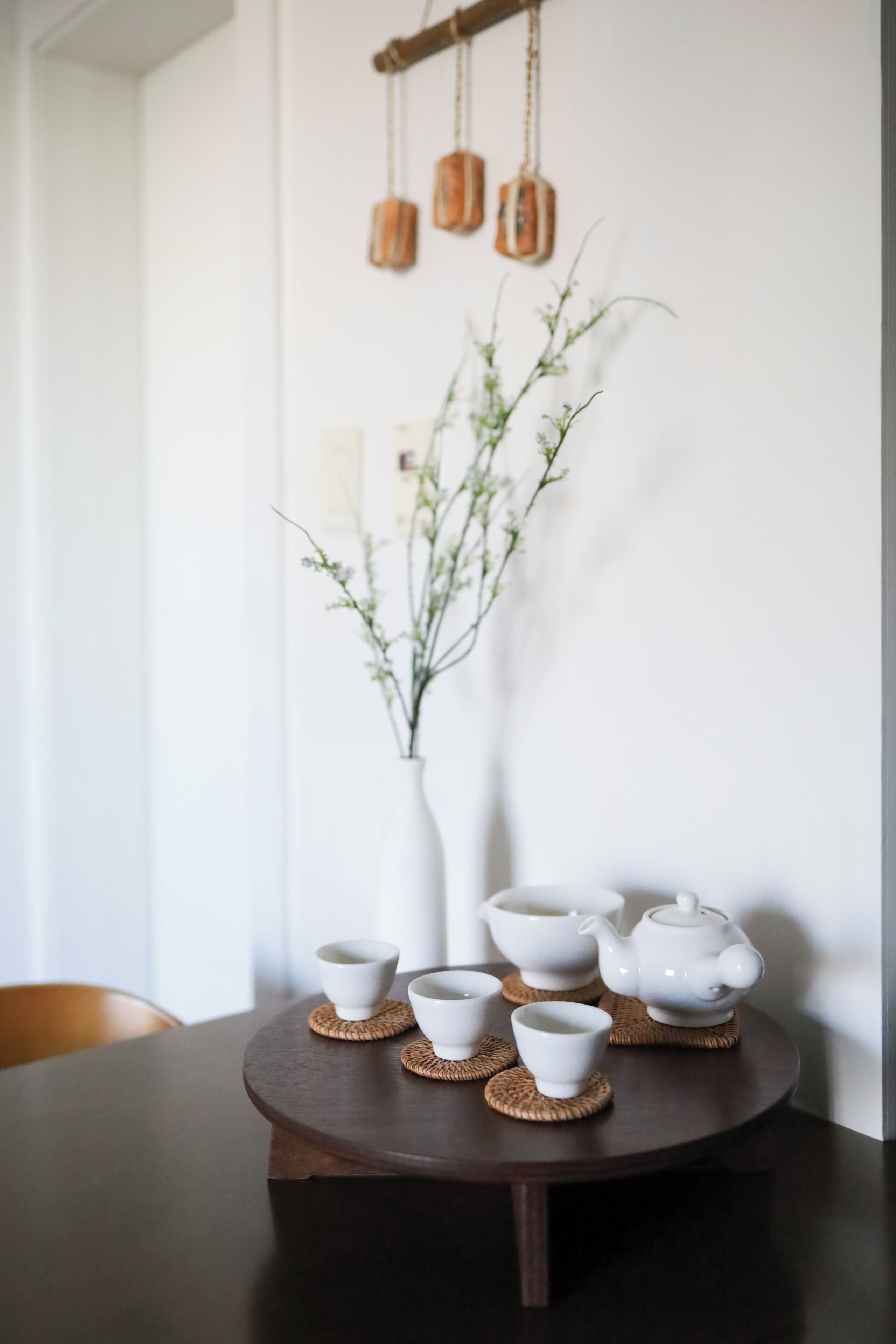 A round wooden table with white teacups and a teapot on woven coasters, a white vase with a plant, and hanging copper containers.