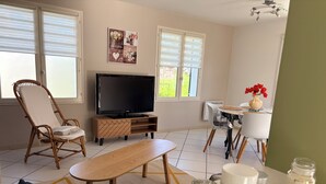 A living room with a TV, wicker chair, dining table, and a small kitchen area.