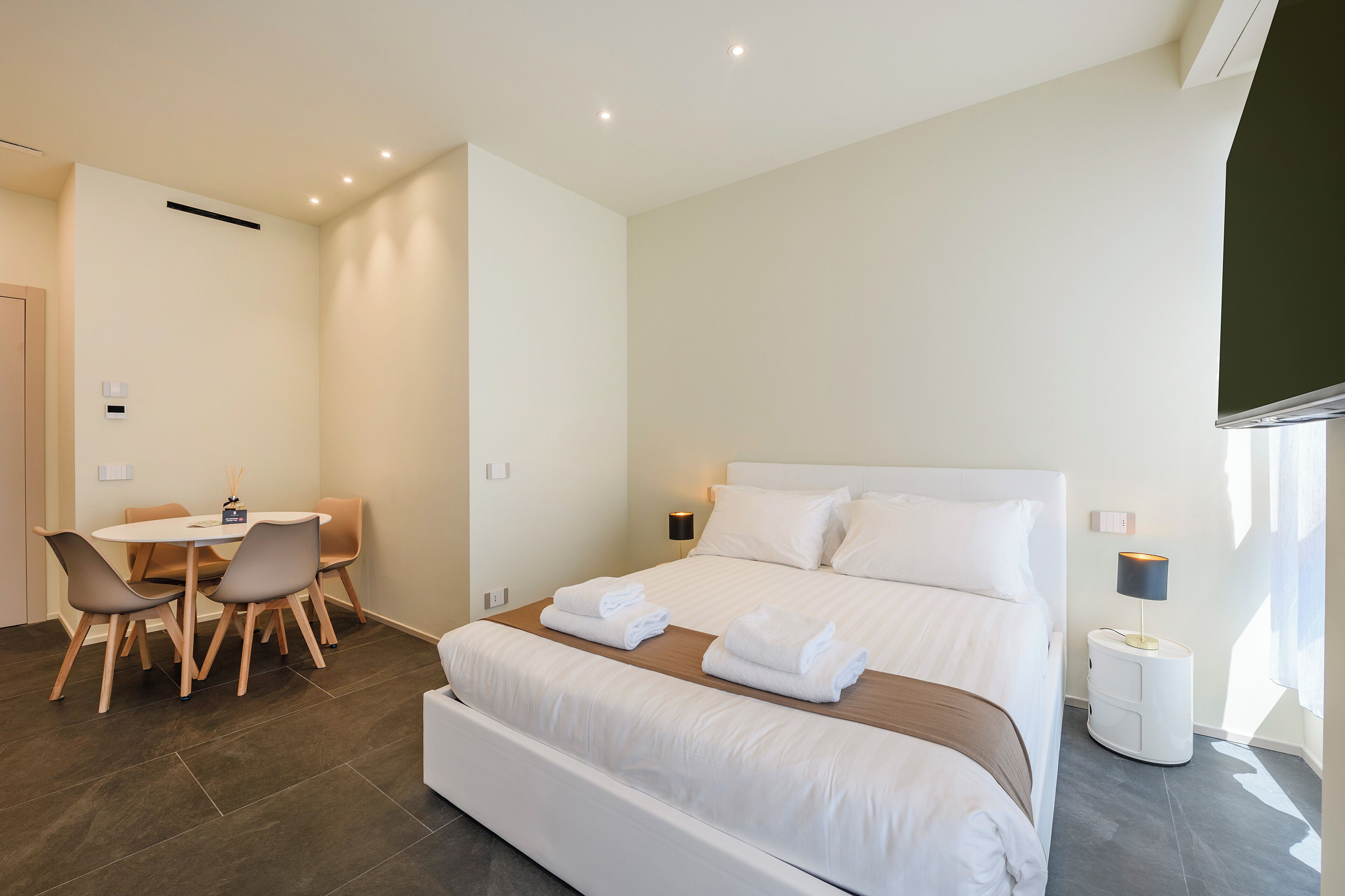 A hotel room with a bed, a desk with chairs, and a bedside table with a lamp.