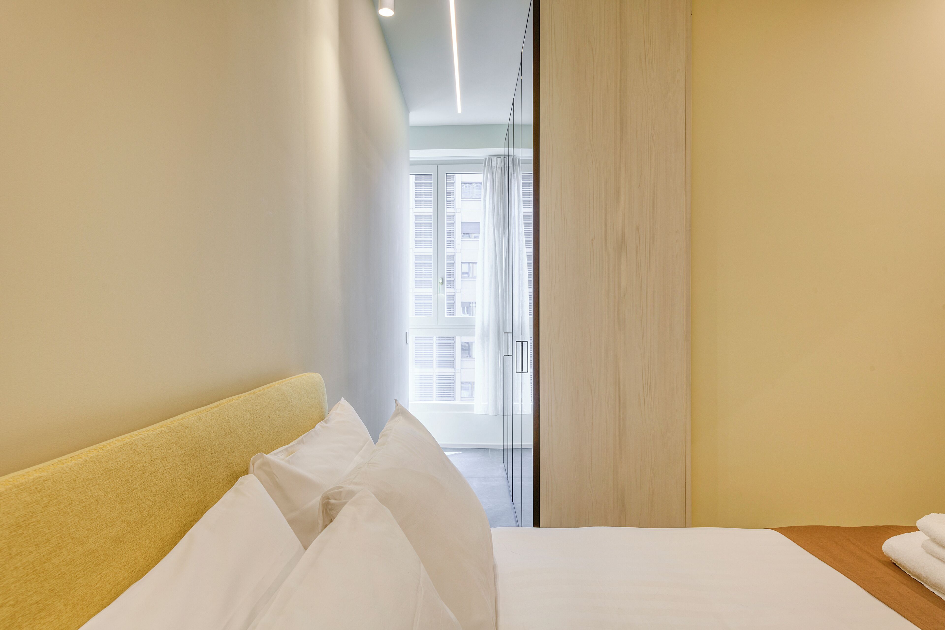 Habitación de hotel con cama, cabecera de madera, ventana con cortinas y vista a un edificio.