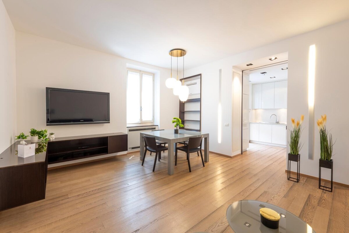 A modern living room with a flat-screen TV, a dining area with a glass table, and a kitchen in the background.