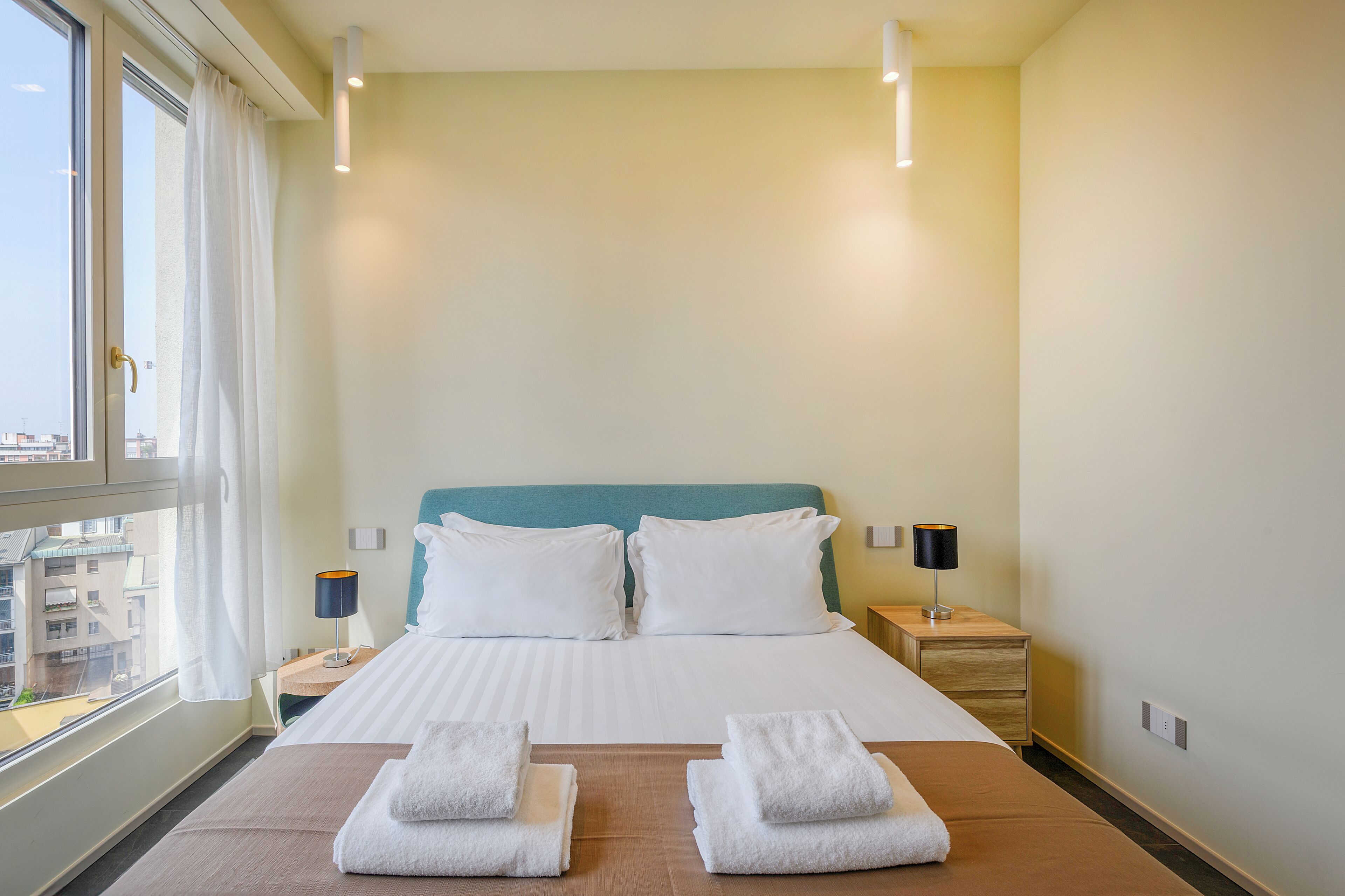 A hotel room with a bed, two bedside tables, a window with curtains, and a view of buildings outside.