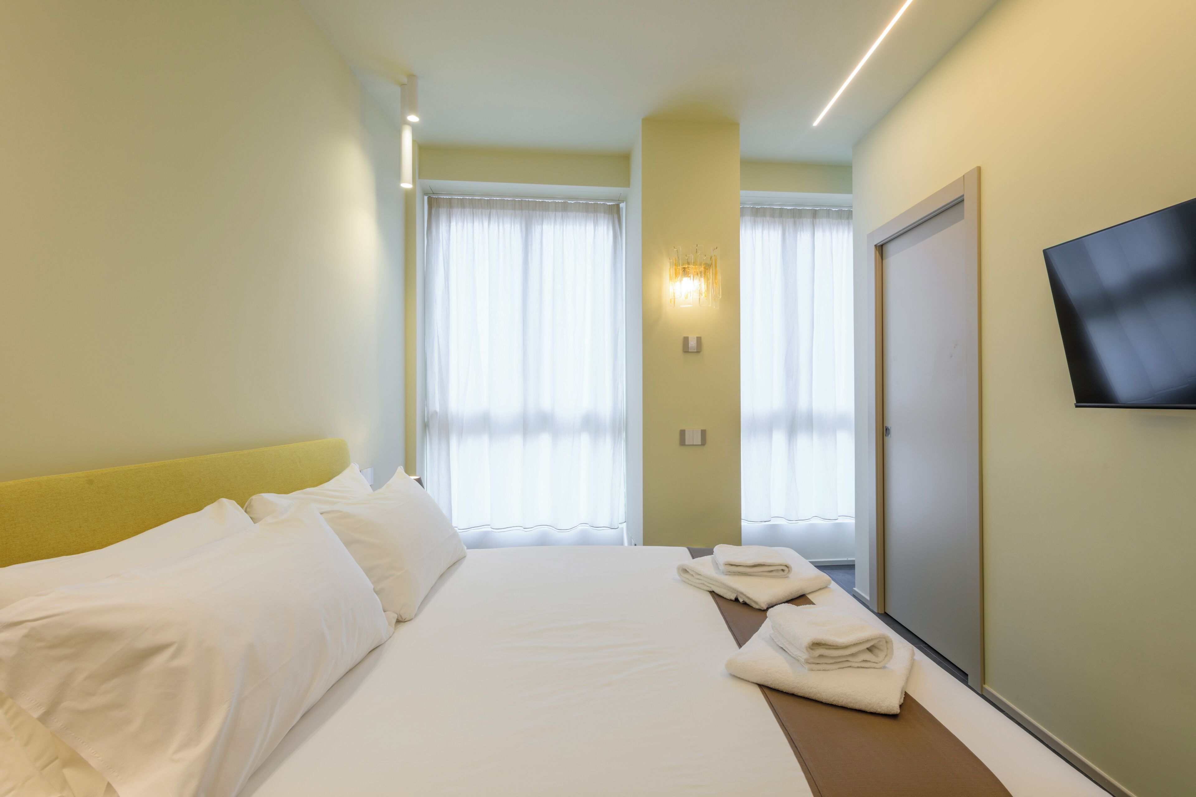 Habitación de hotel con cama, televisor montado en la pared y ventana con cortinas.